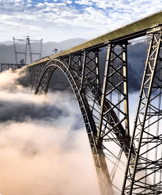 View of Chenab Rail Bridge, a tall steel arch bridge spanning dramatic mist-covered mountains and valleys